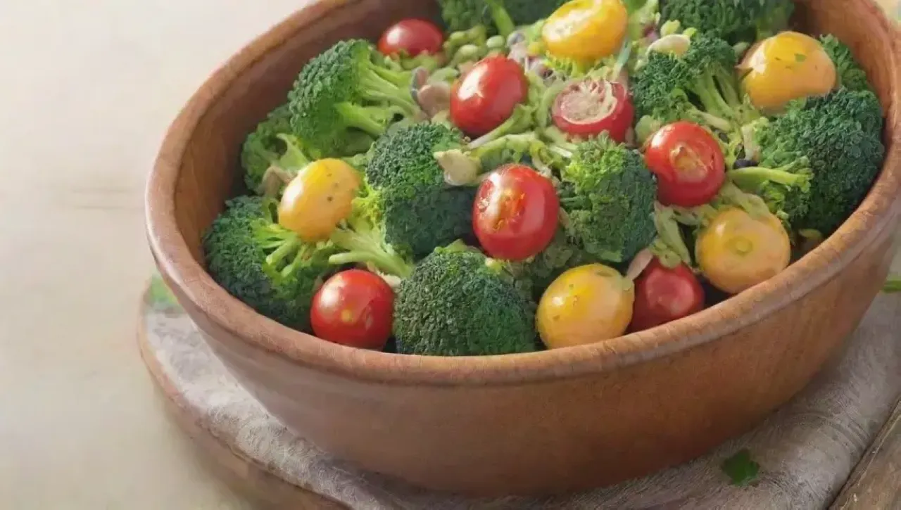 En un tazón rústico, se presenta una ensalada colorida y fresca con brócoli, semillas de girasol, aderezo de mostaza, tomates cherry, cebolla roja, sal marina, perejil y un toque de limón