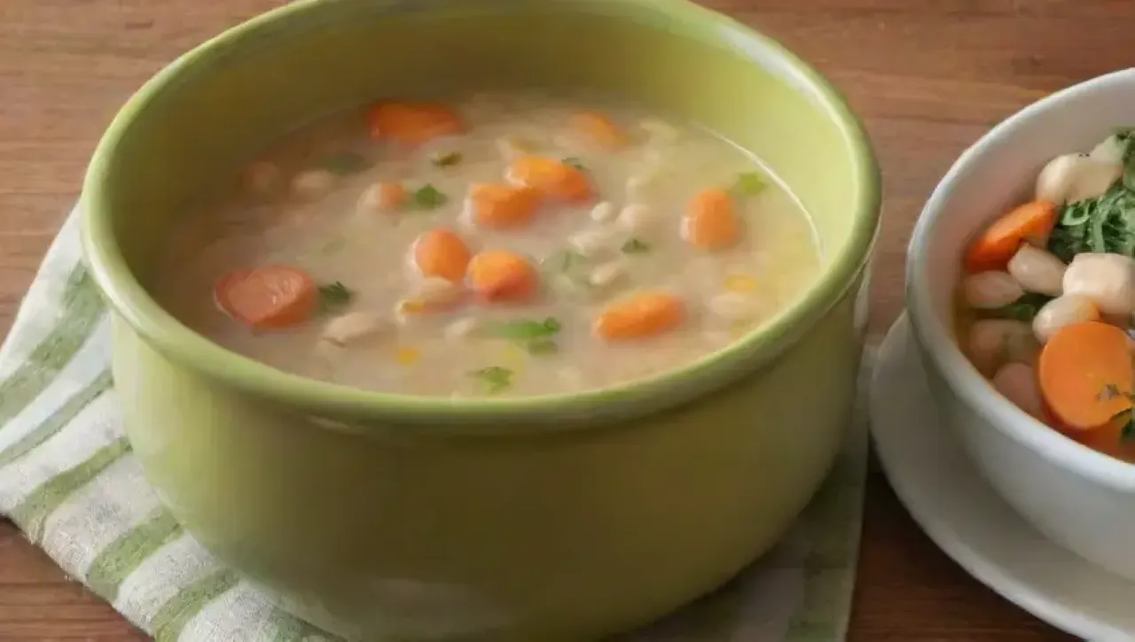 Sopa de frijoles blancos humeante en un tazón cerámico azul, con hierbas frescas, zanahorias, cebollas y ajo, acompañada de pan y un ambiente acogedor